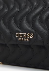 Bolso de cuero negro acolchado que presenta un logo metálico dorado que dice "GUESS EST 1981", con un patrón texturizado y costuras detalladas.