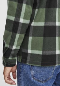 Groene en zwarte geruite flanellen blouse met een zachte textuur; heeft een knoopmanchet en wordt gedragen met blauwe denim jeans.