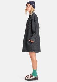Gestreiftes Hemdkleid aus schwarz-weißem Stoff mit langen Ärmeln, einer Tasche und lockerer Passform, kombiniert mit gemusterten Schuhen und grünen Socken.