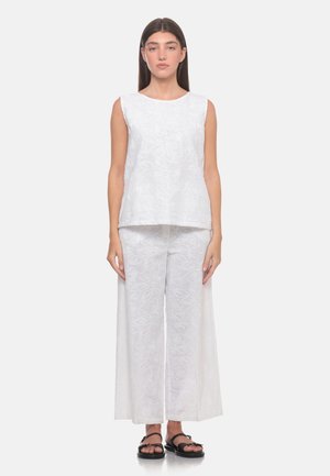 Donna in piedi che indossa una canotta bianca senza maniche e pantaloni a gamba larga con sottili motivi floreali e sandali neri bassi su sfondo bianco.
