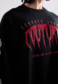 Yli suuri musta t-paita, jossa on punainen graafinen teksti, joka sisältää kuluneen fontin ja lisätekstiä alapuolella. Koottu puuvillasta, jonka pinta on sileä.