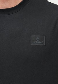 Μαύρο πουκάμισο με στρογγυλή λαιμόκοψη και μια μικρή μαύρη ορθογώνια ετικέτα με το λογότυπο Timberland στο αριστερό στήθος.