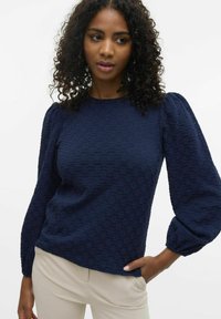Maglietta lunga navy con texture, maniche a sbuffo e scollatura rotonda, abbinata a pantaloni beige.