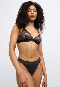 Μαύρο σουτιέν από δαντέλα με τριγωνικό σχήμα κύπελλου και ρυθμιζόμενους ιμάντες. Συνδυάζεται με τανγκό εσώρουχο από δαντέλα και ύφασμα σε μαύρο χρώμα.