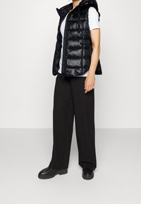 Czarna puchowa kamizelka z błyszczącą fakturą, kapturem i zamkiem błyskawicznym z przodu, noszona na białej koszulce i zestawiona z luźnymi czarnymi spodniami oraz masywnymi butami.