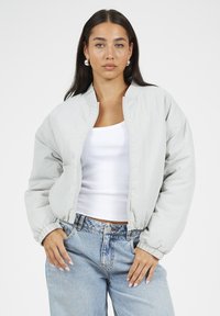 Jasnoszara kurtka bomber z zamkiem z przodu, ściągaczowymi mankietami i luźnym krojem. W połączeniu z białym topem na ramiączkach oraz wyblakłymi niebieskimi dżinsami.