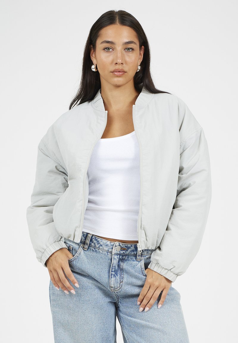 Jasnoszara kurtka bomber z zamkiem z przodu, ściągaczowymi mankietami i luźnym krojem. W połączeniu z białym topem na ramiączkach oraz wyblakłymi niebieskimi dżinsami.