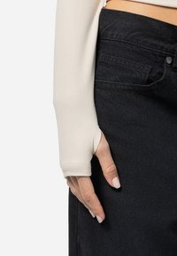 Dlhý rukáv, priliehavý top vo svetlo béžovej tkanine s detailom na palec, v kombinácii s čiernymi denimovými džínami. Textúra vyzerá hladko a elasticky.