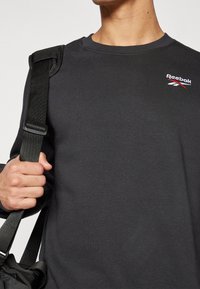 Černá mikina se kulatým výstřihem, logo Reebok v červené a bílé barvě, s texturovanou tkaninou. Popruh od tašky viditelný na rameni.