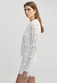 Bílá, průsvitná krajková košile s dlouhými rukávy a límcem; má složité vzory a přiléhavý střih. Vytváří elegantní kombinaci s ozdobnými bílými šortkami.