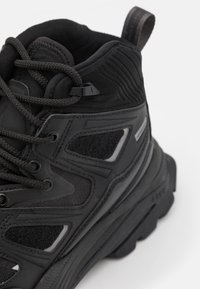 Bota de senderismo negra con tejido texturizado y superposiciones sintéticas, que presenta acentos de goma, cordones estructurados y una suela acolchada.