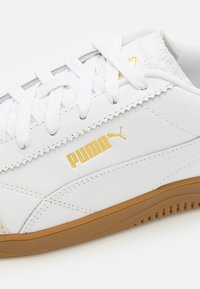 Baskets en cuir blanc avec un logo "PUMA" doré, des accents texturés et une semelle en caoutchouc gomme, lacets plats blancs et design à tige basse.