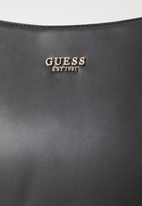 Sort kunstlæderprodukt med et guld-tone "GUESS EST 1981" logo, glat tekstur og minimalistisk design med kontrasterende syninger.
