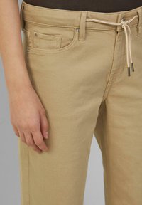 Beige bombažne hlače z elastičnim pasom, z sprednjimi žepi in gladko teksturo. Close-up roke, ki počiva na tkanini.