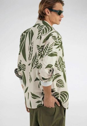 Mann trägt weißes Hemd mit grünem Blattmuster, grüne Hose und Sonnenbrille, steht mit einer Hand in der Tasche und schaut seitlich.