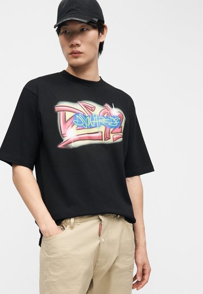 Νεαρός με μαύρο καπέλο μπέιζμπολ, μαύρο T-shirt με πολύχρωμο σχέδιο σε στυλ γκράφιτι και μπεζ φθαρμένο παντελόνι, με το χέρι στην τσέπη.