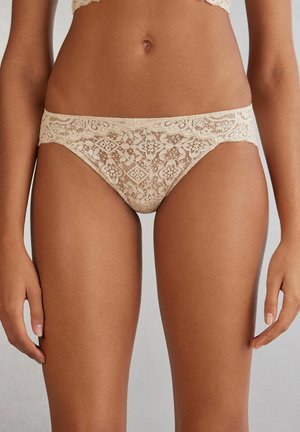 Culotte en dentelle beige avec des motifs floraux, dotée d'une taille ondulée et d'une finition douce et texturée. Conçue pour un ajustement taille basse.