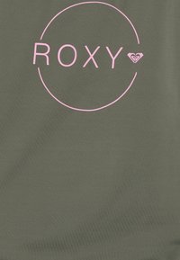 Olivgrön T-shirt med en stor rosa "ROXY"-logotyp i en cirkel, med ett hjärtemblem. Slät textur och avslappnad design.