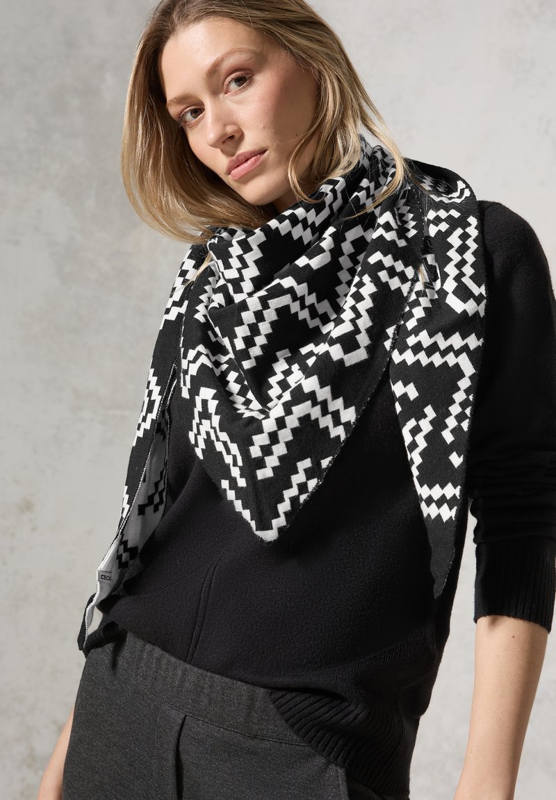 Foulard triangolare a righe bianche e nere realizzato in tessuto morbido, con design a zigzag e orli sfrangiati, indossato sopra un maglione scuro.