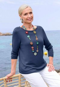 Dunkelblauer Pullover mit Rundhalsausschnitt und Dreiviertelärmeln, kombiniert mit einer bunten, floralen Perlenkette, weißen Hosen, sitzend auf einem Korbstuhl.