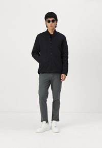 Svart zip-up jakke med høyt krage, grå slim-fit bukser og hvite sneakers. Runde solbriller fullfører antrekket. Jevn tekstur totalt.