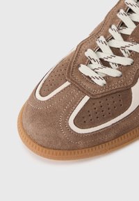 Zapatillas de ante marrón con perforaciones, detalles en blanco y cordones de beige claro. Presentan una suela de goma con patrones texturizados.