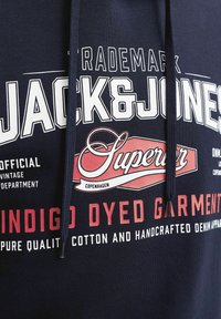 Sweat à capuche bleu marine avec texte blanc et rouge indiquant Jack & Jones, Superior Copenhagen, vêtement teint à l'indigo, coton de pure qualité et vêtements en denim réalisés à la main.