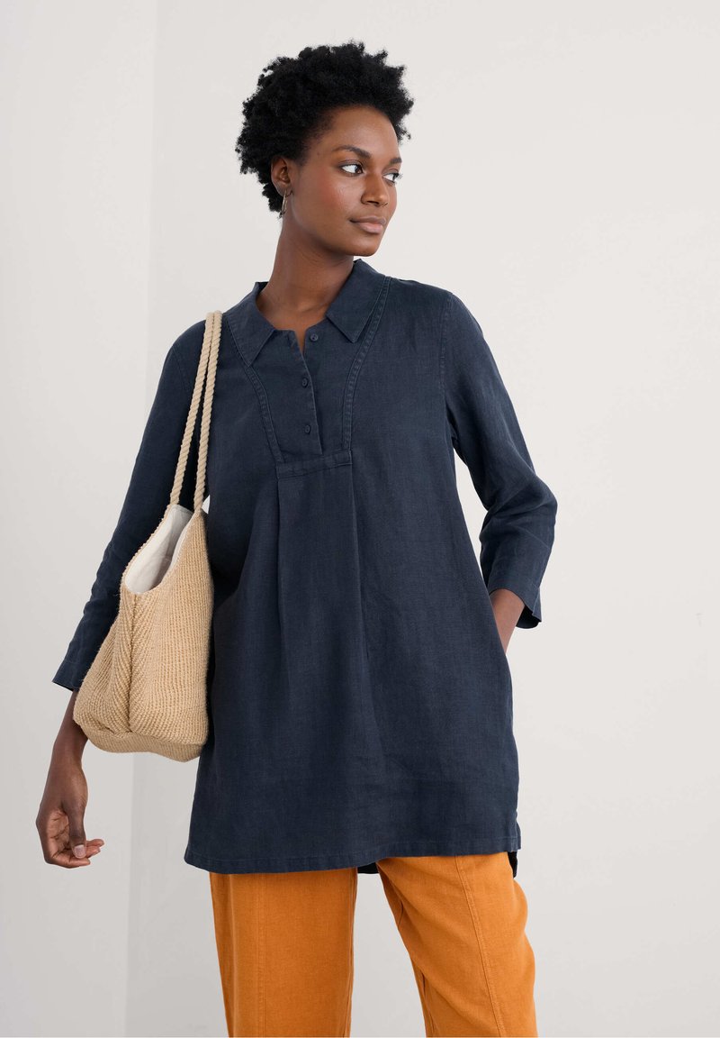 Marineblauwe linnen tuniek met een kraag, knoopsluiting en plooidetail. Gepaard met oranje broeken en een gestructureerde beige schoudertas.