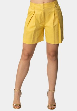 Donna che indossa pantaloncini gialli plissettati a vita alta e sandali dorati con tacco alto e cinturino alla caviglia, in piedi davanti a uno sfondo neutro.