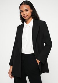 Černé oversized sako s špičatými klopami, se dvěma předními knoflíky, nošené přes bílou košili s límečkem, doplněné černými strukturovanými kalhotami.