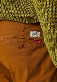 Pantalon en coton marron avec un pull en maille verte texturée partiellement visible. Les caractéristiques incluent une fermeture à boutons, une poche cousue et une étiquette Levi's.
