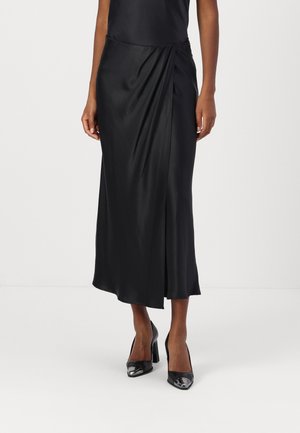 Femme portant une jupe midi en satin noir avec un détail portefeuille asymétrique et des chaussures à talons hauts en cuir verni noir, debout devant un fond blanc.