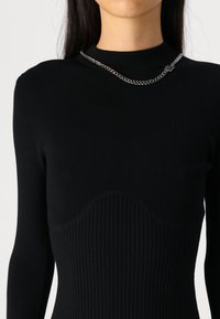 Svart ribbestrikket topp med lange ermer og høy hals, med en buet designdetalj ved brystet og en sølvkjede.