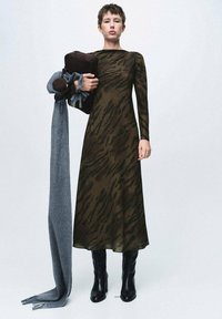 Vestido de manga larga en verde oliva oscuro con un patrón de rayas de tigre, combinado con una bufanda gris y botas negras hasta la rodilla, sostenido con un bolso marrón.