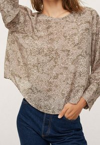 Blouse légère à motifs beige avec des taches sombres, col rond et manches longues. Le tissu est transparent avec une coupe ample et fluide.