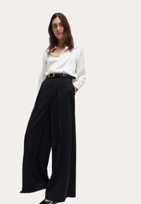 Camisa blanca de botones metida dentro de unos pantalones negros de pierna ancha y cintura alta, asegurados con un cinturón negro con hebilla de oro.