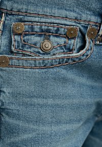 Gros plan d'une poche de jean en denim bleu avec couture marron et cinq rivets en laiton, dont un avec logo de marque embossé et texte.