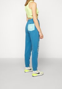 Blue sweatpants with a mint green patch pocket, elastic waistband, printed "SILVER" logo, and paired with white and black sneakers.