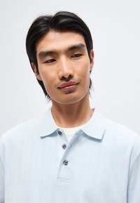 Νέος άνδρας με μαύρα μαλλιά, φοράει ένα ανοιχτομπλε πόλο πουκάμισο με κουμπιά, κοιτάζει ελαφρώς προς τα κάτω με φόντο ένα απλό λευκό φόντο.