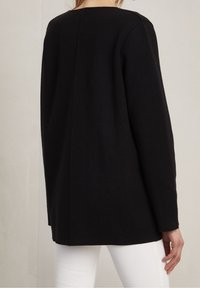 Black, long-sleeve sweater with a straight cut and back seam detail, made of a textured fabric. White pants visible in the lower part.