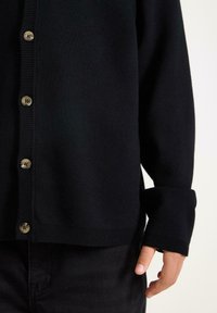 Černý pletený cardigan s knoflíkovým zapínáním vpředu, zdobený hnědými knoflíky a žebrovanými manžetami. Měkká textura, volný střih.