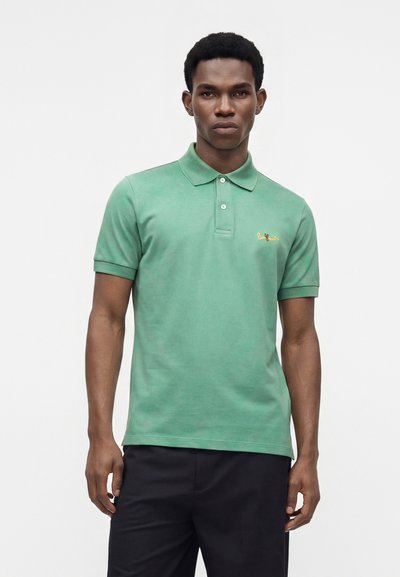 Homme portant un polo vert à manches courtes avec col et un pantalon noir, debout devant un fond clair uni.