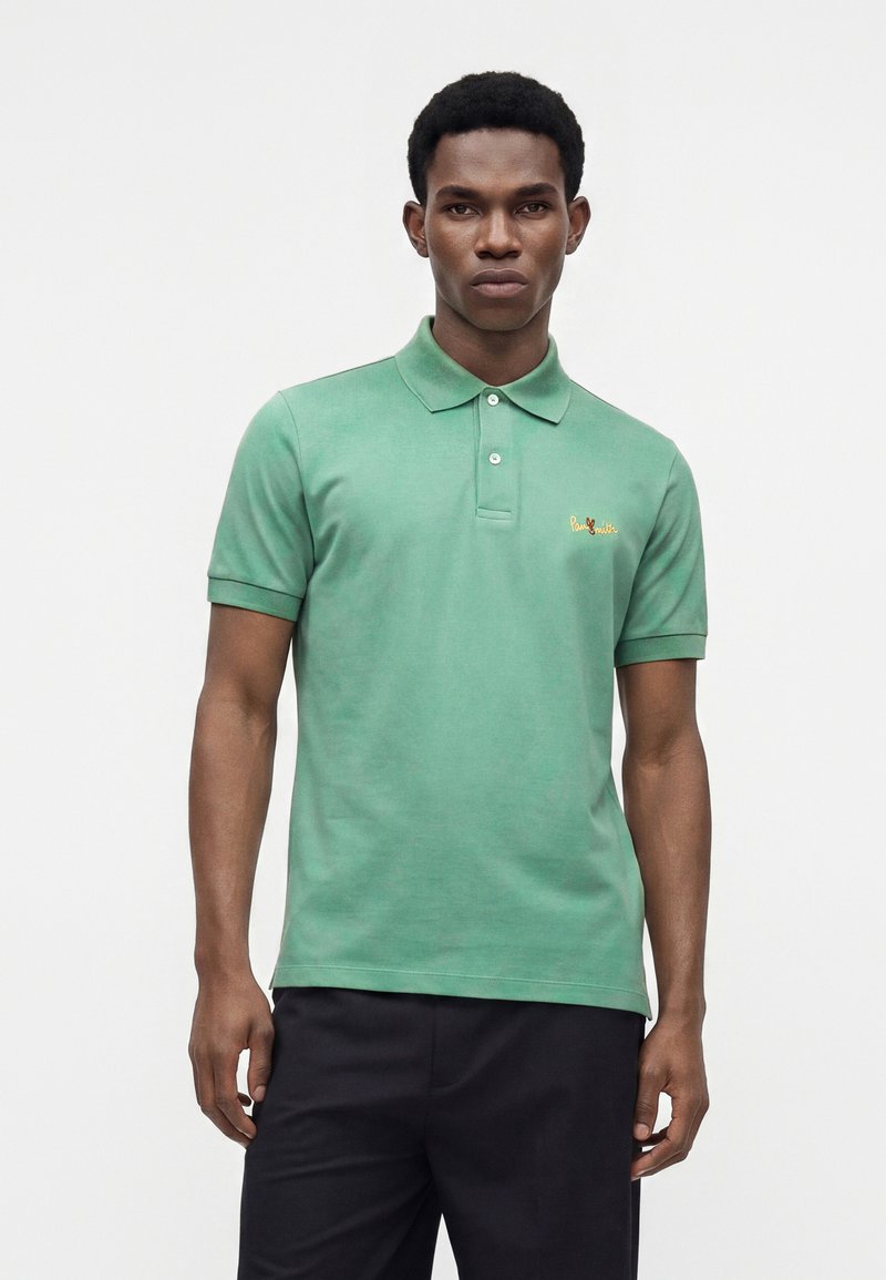 Homme portant un polo vert à manches courtes avec col et un pantalon noir, debout devant un fond clair uni.