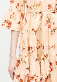 Φόρεμα σε ανοιχτό μπεζ με floral σχέδιο, μανίκια καμπάνα, φούστα σε στρώσεις και ζώνη στη μέση με κόκκινα και πορτοκαλί λουλούδια.