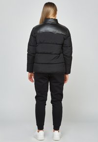 Svart puffjacka med en glänsande svart axelremsa, quiltat mönster och hög krage. Tillsammans med svarta mjukisbyxor och vita sneakers.