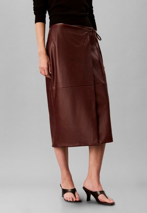 Femme portant une jupe midi portefeuille en cuir marron avec un lien à la taille et des sandales à talons noires à bout ouvert, debout devant un fond gris.