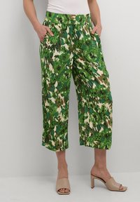 Pantaloni a gamba larga verdi e crema con un motivo floreale, dotati di due tasche laterali e una texture liscia, abbinati a sandali a scivolo color nude.