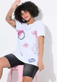 T-shirt blanche avec des imprimés graphiques colorés, comprenant des phrases et des étoiles. Associée à un short en denim foncé avec des bords effilochés.