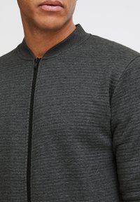 Chaqueta bomber gris oscuro con un tejido texturizado, cuello alzado y cremallera frontal. Presenta un diseño ajustado sin logotipos ni adornos visibles.