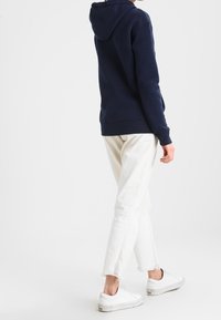 Person som bär en marinblå hoodie, off-white korta jeans med fransade kanter och vita sneakers, stående mot en enkel ljus bakgrund.